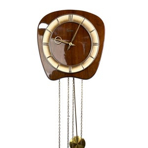 May include: A wall clock with a dark brown wooden frame, a circular face featuring a cream-coloured ring with gold-tone markers, and gold hands. It includes three hanging chains, two cylindrical brass weights, and a circular brass pendulum.
