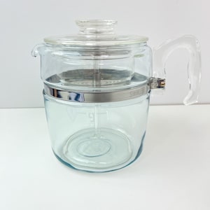 May include: A clear glass coffee percolator with a stainless steel band and a glass lid. The percolator has a clear glass handle.
