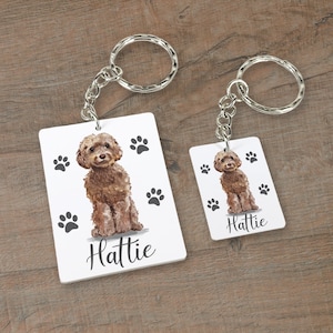 May include: Two keychains with a brown dog illustration and the name "Hattie" printed on each. The keychains are on a wooden surface.