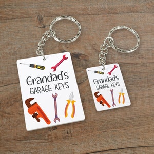 May include: Two white keychains with silver key rings. The keychains are rectangular and feature a white background with a red wrench, a black screwdriver, a wrench, and pliers. The text "Grandad's Garage Keys" is printed in black on both keychains.