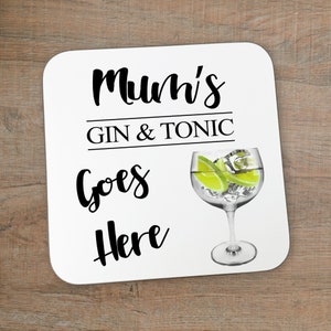 Puede incluir: Posavasos cuadrado blanco con texto negro que dice "Mum's Gin & Tonic Goes Here". A la derecha del posavasos se ve un vaso de gin tonic con rodajas de lima y hielo.