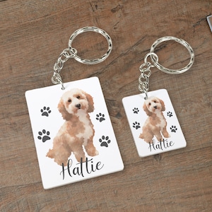 May include: Two rectangular keychains with silver rings. Each keychain features a watercolor illustration of a light brown dog with black paw prints and the name "Hattie" in cursive. The keychains are on a wooden surface.