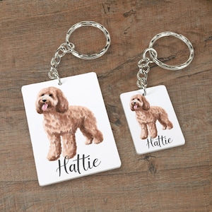 May include: Two keychains featuring a watercolor illustration of a brown doodle dog. The larger keychain has the name "Hattie" printed below the dog. The smaller keychain has the name "Hattie" printed below the dog.