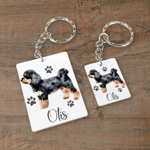 May include: Two white rectangular keychains with black paw prints and the name "Otis" printed on them. Each keychain features a watercolor painting of a black and brown dog.