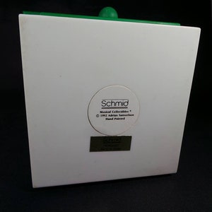 May include: White ceramic figurine with a green top. The figurine has a label on the back that reads "Schmid Musical Collectibles &copy; 1992 Adrian Samourkash Hand Painted".