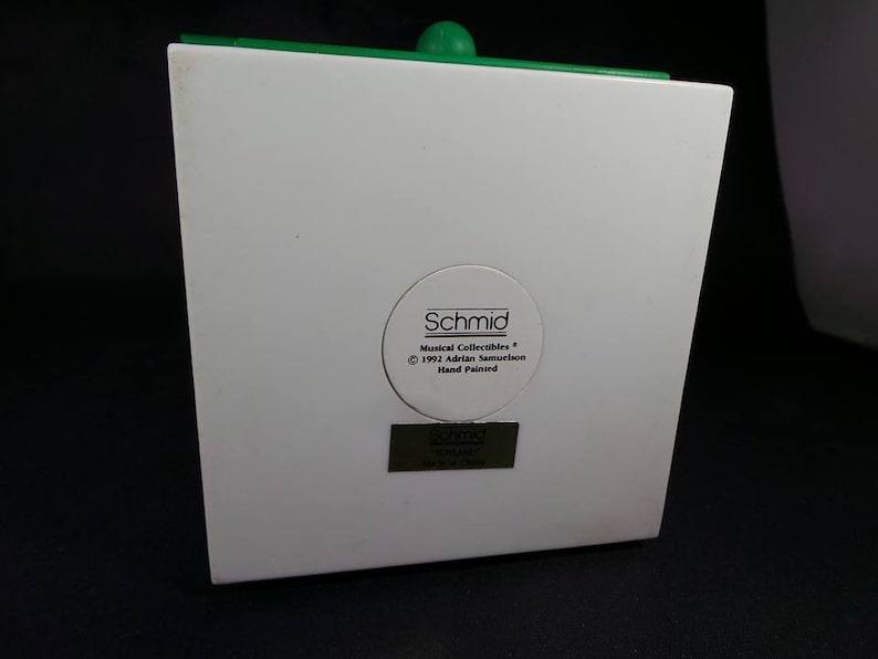 May include: White ceramic figurine with a green top. The figurine has a label on the back that reads "Schmid Musical Collectibles &copy; 1992 Adrian Samourkash Hand Painted".