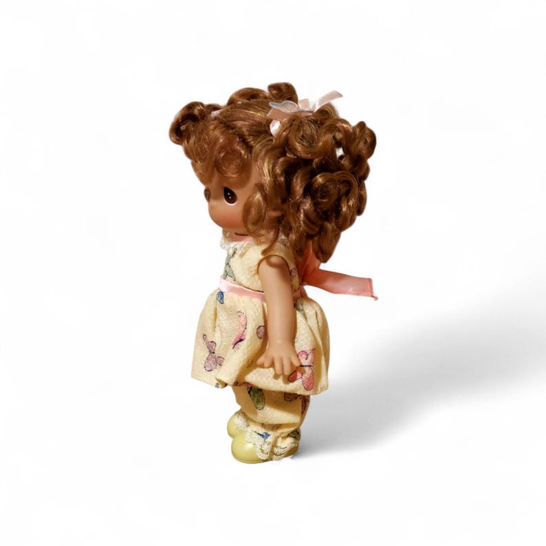 May include: A small doll with brown curly hair, wearing a yellow dress and pants with a butterfly pattern. The doll has a pink ribbon and a white bow in its hair. The doll is standing on a white surface.