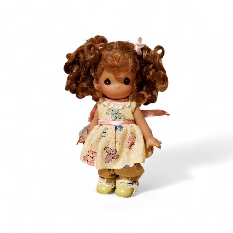 May include: A collectible doll with brown curly hair styled in pigtails, wearing a yellow dress and pants adorned with butterfly designs. The doll has a sweet expression and is holding a small object. The outfit is accented with pink ribbons and light green shoes.