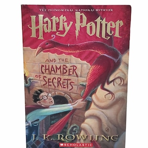 May include: A red, white, and gold book cover with the title "Harry Potter and the Chamber of Secrets" by J.K. Rowling. The cover features an illustration of Harry Potter reaching for a red phoenix.