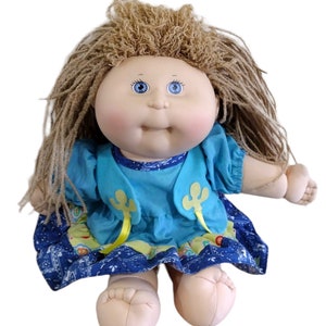 May include: A Cabbage Patch Kid doll with long brown hair, blue eyes, and a blue and yellow dress. The doll is sitting with its legs crossed.