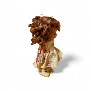 May include: A vintage doll with brown curly hair, wearing a yellow dress with butterfly patterns and a pink ribbon. The doll has a sweet expression and is standing on a white surface.