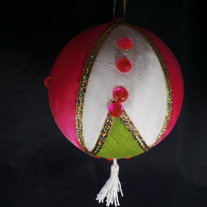 Puede incluir: Un adorno navide&ntilde;o de color rosa, blanco y verde con un borla blanca. El adorno est&aacute; decorado con purpurina rosa y cinta dorada.