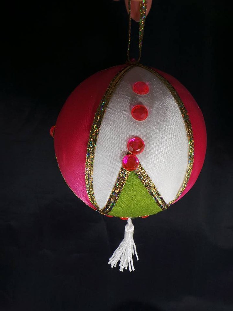 Puede incluir: Un adorno navide&ntilde;o de color rosa, blanco y verde con un borla blanca. El adorno est&aacute; decorado con purpurina rosa y cinta dorada.