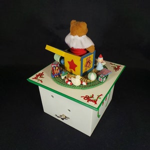 May include: A white wooden music box with a green trim. The top of the box features a scene of a teddy bear popping out of a toy box with other toys around it. The music box has a winding key on the bottom.