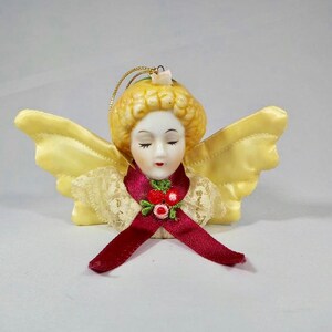 Può includere: Un ornamento a forma di angelo in ceramica con un volto sereno, occhi chiusi e capelli biondi. Presenta ali in raso giallo, un nastro bordeaux e un colletto in pizzo. L'ornamento è adornato con piccoli accenti floreali rossi e un anello dorato per appenderlo.