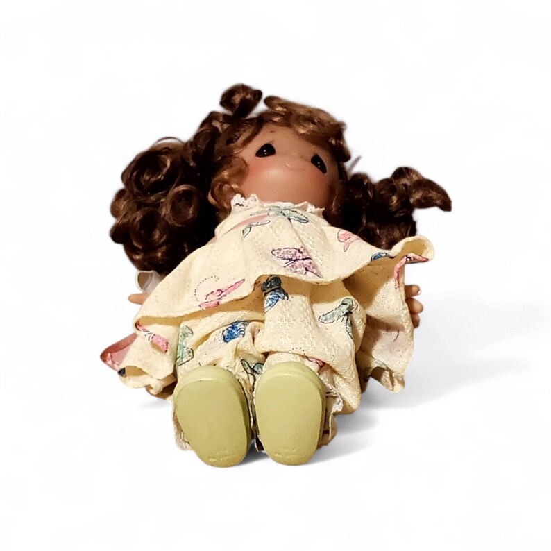 May include: A vintage doll with brown curly hair and a painted face. The doll wears a cream-colored dress and matching pants with a butterfly pattern. The doll's shoes are light green.