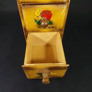 May include: A wooden box with three drawers. Each drawer has a painted design and a label. The top drawer says "Buttons", the middle drawer says "Needles & Pins", and the bottom drawer says "Needles".