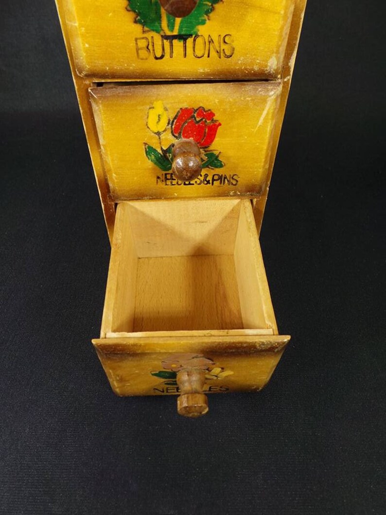 May include: A wooden box with three drawers. Each drawer has a painted design and a label. The top drawer says "Buttons", the middle drawer says "Needles & Pins", and the bottom drawer says "Needles".