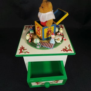 May include: A wooden music box with a green drawer and a white top. The top features a teddy bear popping out of a toy box with ABC blocks and other toys. The music box is decorated with holly and red ribbons.