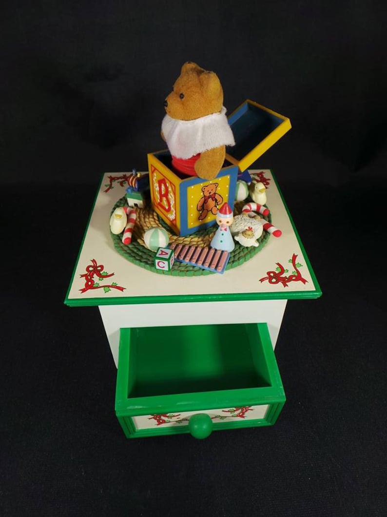 May include: A wooden music box with a green drawer and a white top. The top features a teddy bear popping out of a toy box with ABC blocks and other toys. The music box is decorated with holly and red ribbons.