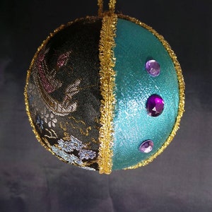 Puede incluir: Un adorno navide&ntilde;o hecho a mano con un exterior de tela negro y turquesa. El adorno est&aacute; decorado con ribetes dorados y diamantes de imitaci&oacute;n morados.