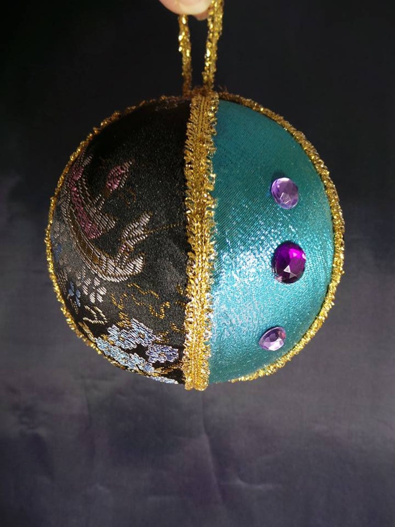 Puede incluir: Un adorno navide&ntilde;o hecho a mano con un exterior de tela negro y turquesa. El adorno est&aacute; decorado con ribetes dorados y diamantes de imitaci&oacute;n morados.