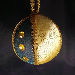 Puede incluir: Un adorno navide&ntilde;o dorado y azul verdoso con ribete dorado y diamantes de imitaci&oacute;n. El adorno es redondo y tiene una cuerda dorada atada a la parte superior.