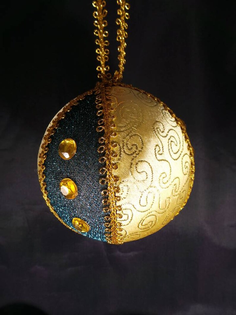 Puede incluir: Un adorno navide&ntilde;o dorado y azul verdoso con ribete dorado y diamantes de imitaci&oacute;n. El adorno es redondo y tiene una cuerda dorada atada a la parte superior.