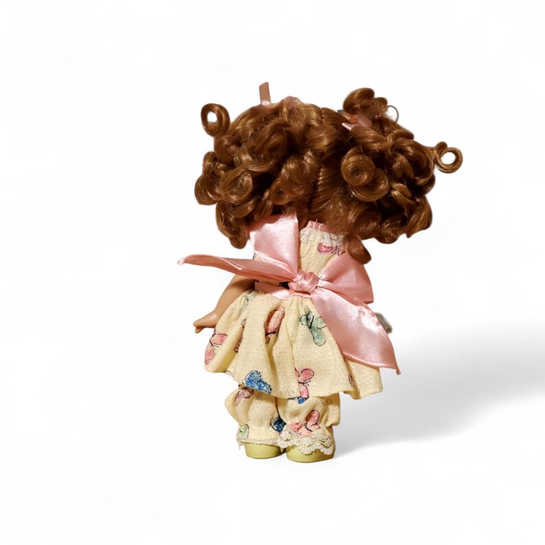 May include: A vintage doll with brown curly hair and a pink bow. The doll wears a yellow dress and pants with a butterfly pattern. The doll's outfit is accented with pink bows.