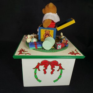 May include: A white wooden music box with a green trim and a red bow. The top of the box features a pop-up toy of a teddy bear in a box with other toys, including a ball, a train, and blocks. The box plays music.