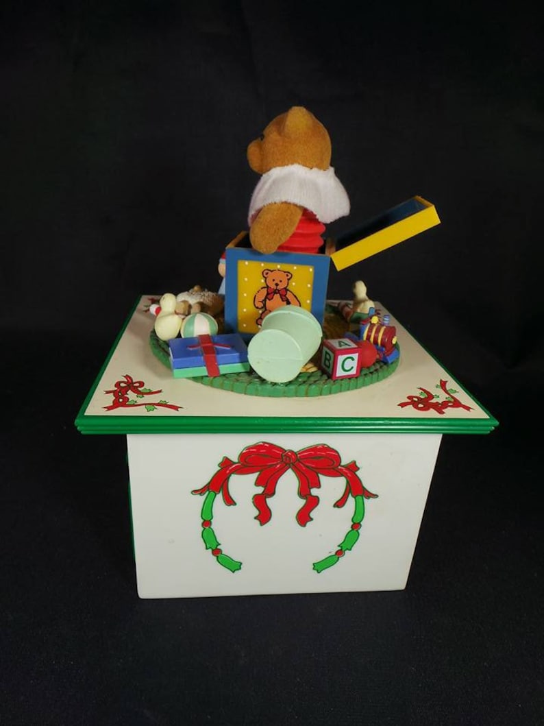 May include: A white wooden music box with a green trim and a red bow. The top of the box features a pop-up toy of a teddy bear in a box with other toys, including a ball, a train, and blocks. The box plays music.
