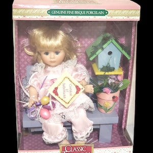 Könnte beinhalten: Eine Porzellanpuppe mit blonden Haaren und einem rosa Kleid sitzt auf einer Bank mit einem Vogelhaus und einem Blumentopf. Die Puppe hält einen Ballonstrauß. Die Schachtel trägt die Aufschrift "Classic Treasures Special Edition Collectible Doll" und "Genuine Fine Bisque Porcelain".
