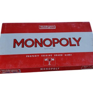 May include: A red and white Monopoly board game box. The box features the word "MONOPOLY" in large red letters, with "PROPERTY TRADING BOARD GAME" printed below. The brand name "Waddingtons" is visible on the box.