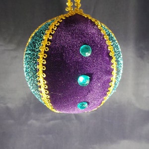 Puede incluir: Adorno navide&ntilde;o de terciopelo morado con ribetes dorados y tres gemas turquesa.
