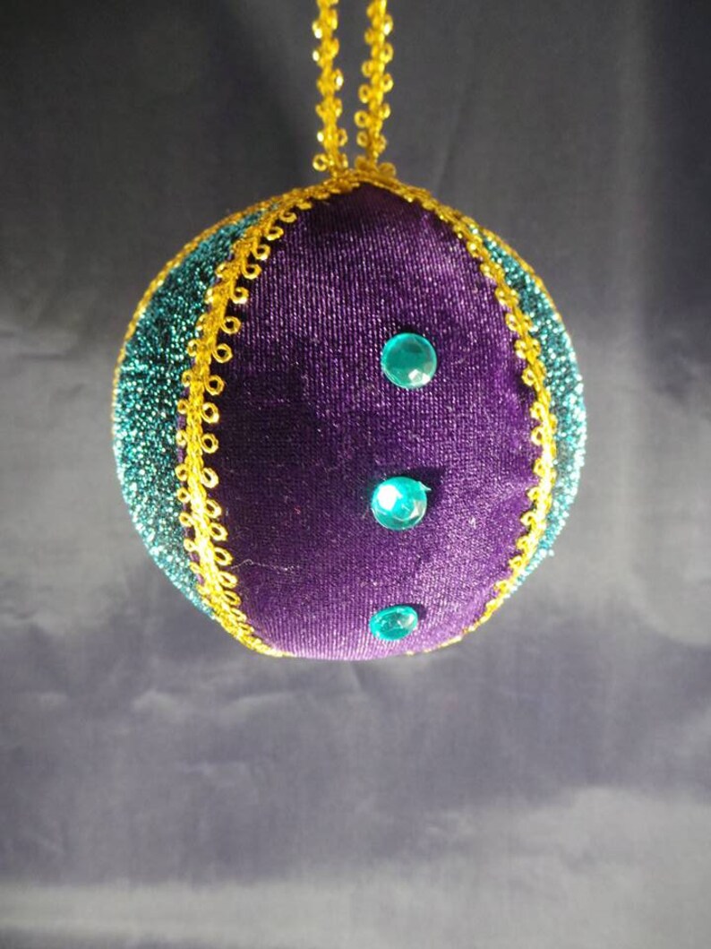 Puede incluir: Adorno navide&ntilde;o de terciopelo morado con ribetes dorados y tres gemas turquesa.