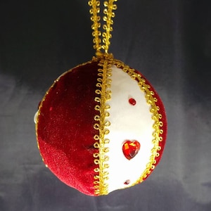 Puede incluir: Un adorno navide&ntilde;o de terciopelo rojo y tela blanca con ribetes dorados y acentos de diamantes de imitaci&oacute;n rojos.