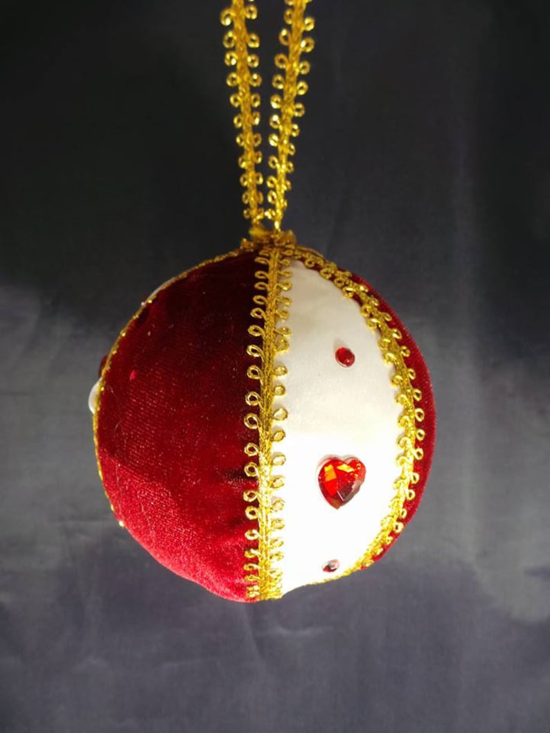 Puede incluir: Un adorno navide&ntilde;o de terciopelo rojo y tela blanca con ribetes dorados y acentos de diamantes de imitaci&oacute;n rojos.