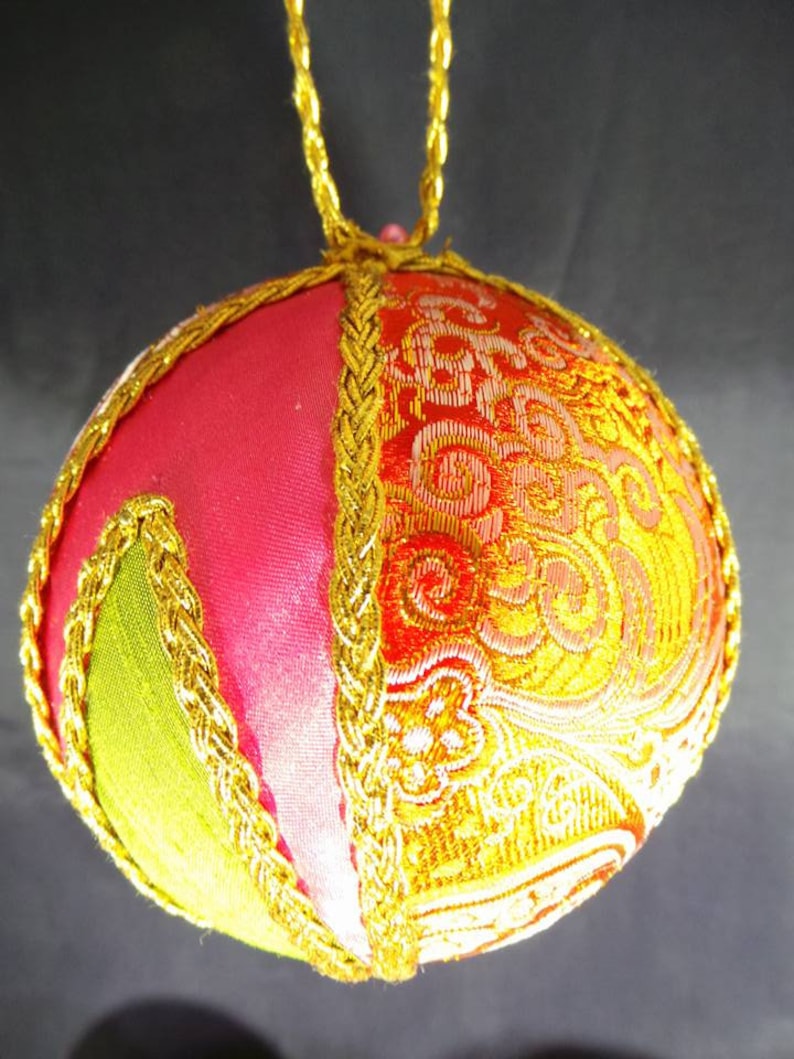Puede incluir: Un adorno navide&ntilde;o hecho a mano en forma de esfera, hecho de tela rosa, verde y dorada con un ribete de trenza dorada. El adorno tiene un dise&ntilde;o tradicional y es perfecto para agregar un toque de elegancia a tu decoraci&oacute;n navide&ntilde;a.