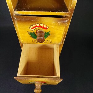 May include: A wooden spice rack with three drawers. The drawers have painted floral designs and the word "BLOOMS" is written on the front of the middle drawer.