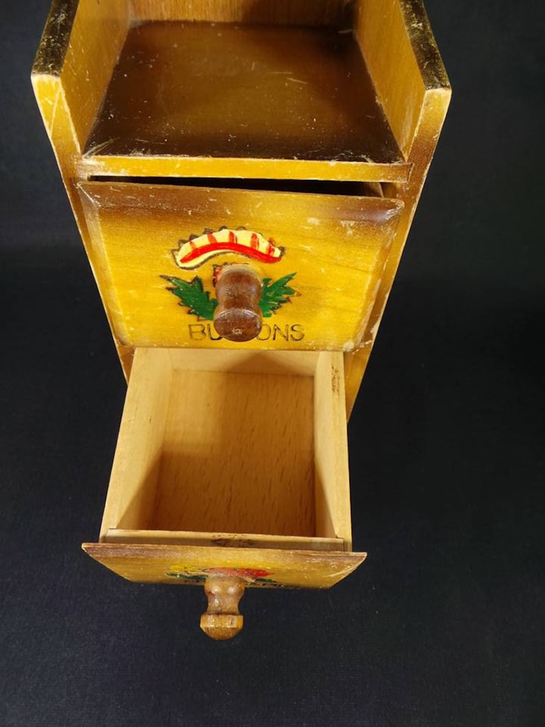 May include: A wooden spice rack with three drawers. The drawers have painted floral designs and the word "BLOOMS" is written on the front of the middle drawer.