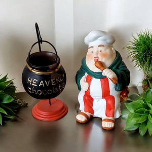May include: A black and gold decorative cauldron with the words "HEAVENLY chocolate" in gold lettering, sitting on a red base. Beside it is a ceramic chef figurine wearing a white hat, green robe, and red and white striped pants.