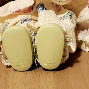 May include: Close-up of a doll's feet, showing the soles of the shoes. The shoes are a pale yellow color and have a rounded shape. The doll's dress is visible, with a floral pattern and lace trim. The background is a wooden surface.