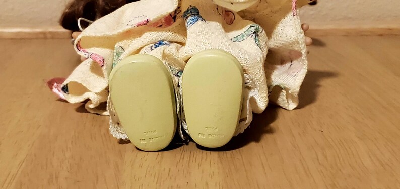 May include: Close-up of a doll's feet, showing the soles of the shoes. The shoes are a pale yellow color and have a rounded shape. The doll's dress is visible, with a floral pattern and lace trim. The background is a wooden surface.