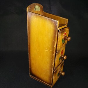 May include: A wooden spice rack with three drawers. Each drawer has a painted floral design and a small wooden knob. The rack is stained a light brown color and has a small label on the top that reads "Just a Pinch of the Good Stuff".
