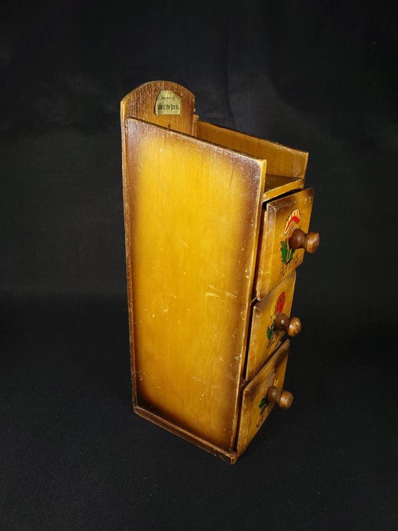May include: A wooden spice rack with three drawers. Each drawer has a painted floral design and a small wooden knob. The rack is stained a light brown color and has a small label on the top that reads "Just a Pinch of the Good Stuff".