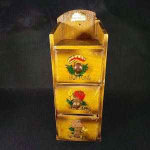 May include: A wooden box with three drawers, each with a different floral design and label. The top drawer is labeled "Buttons", the middle drawer is labeled "Needles & Pins", and the bottom drawer is labeled "Needles".