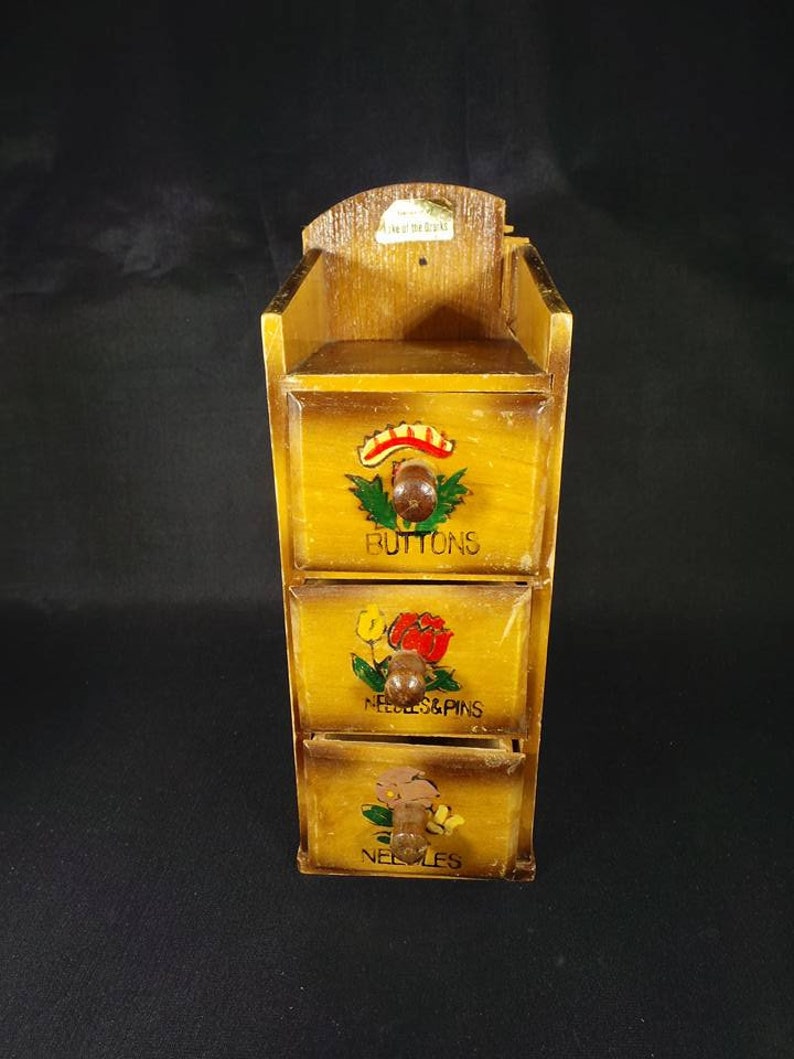 May include: A wooden box with three drawers, each with a different floral design and label. The top drawer is labeled "Buttons", the middle drawer is labeled "Needles & Pins", and the bottom drawer is labeled "Needles".