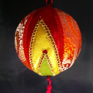 Puede incluir: Un adorno navide&ntilde;o de tela rojo, dorado y verde con un borla. El adorno est&aacute; hecho de diferentes telas y tiene un dise&ntilde;o acolchado.