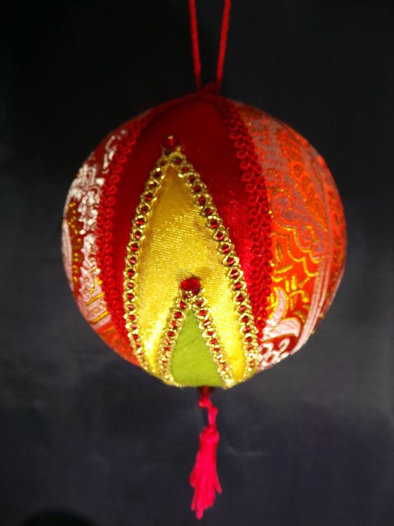 Puede incluir: Un adorno navide&ntilde;o de tela rojo, dorado y verde con un borla. El adorno est&aacute; hecho de diferentes telas y tiene un dise&ntilde;o acolchado.
