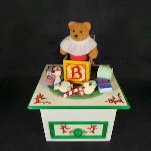 May include: A wooden music box with a teddy bear popping out of a toy block. The box is decorated with a green border and red ribbons. The box has a drawer with a green knob and a green and white design.